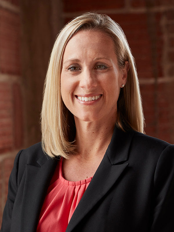 Micaela (Shelton) Banach wearing a coral blouse and black blazer, standing indoors against a warm-toned brick background.