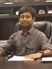 Ravi Atreyapurapu seated at a desk in an office, wearing a striped button‑down shirt, with papers, a pen, and shelving in the background.
