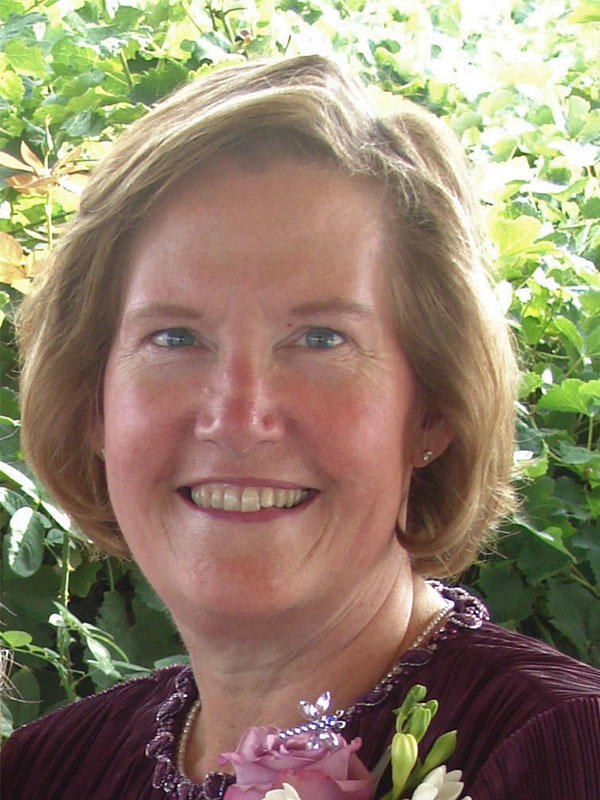 Stacie Hamilton wearing a deep burgundy outfit with a beaded neckline and floral corsage, standing outdoors in front of lush green foliage.
