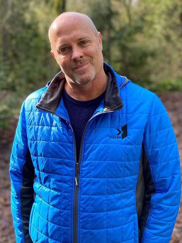 Tim Mulligan wearing a bright blue quilted Vulcan jacket and navy shirt, standing on a wooded trail with soft greenery in the background.