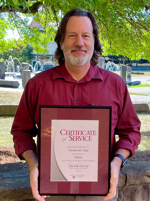 Tom Tripp holding a framed 30‑year Certificate of Service from Washington State University, standing outdoors near fountains and greenery.