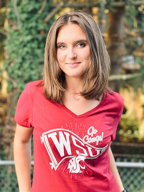 Vanessa Avdeyeva wearing a red WSU ‘Go Cougs!’ T‑shirt, standing outdoors in front of trees with sunlight filtering through the background.