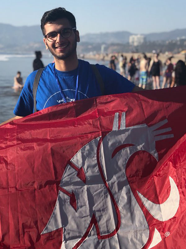Yatin Singla holding a large crimson WSU flag on a sunny beach, with waves, shoreline, and people in the background.