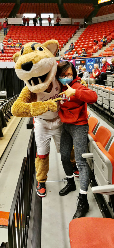 Demi Deng poses with Butch the Cougar mascot in a sports arena, both making playful hand gestures near the stands.