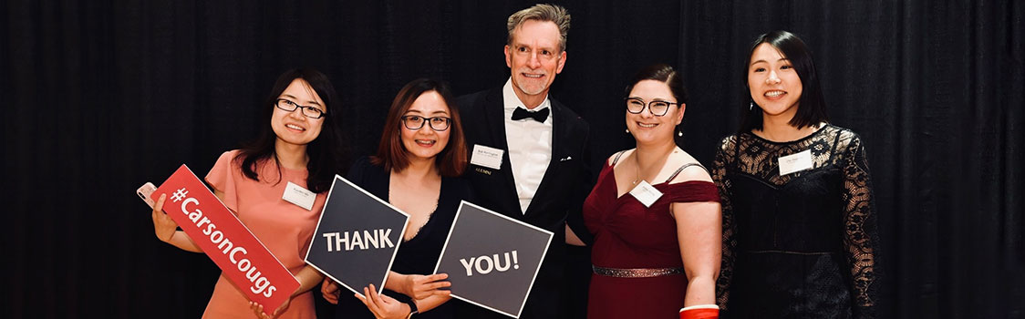 Demi Deng with Dr. Bob Harrington and fellow PhD students posing together during Hospitality Week, holding signs that read “#CarsonCougs” and “Thank you!”