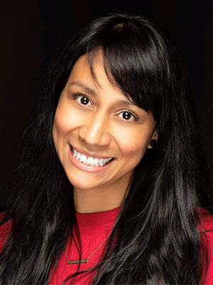Goretti Manzo wearing a red top and a small necklace, with long wavy black hair against a dark background.