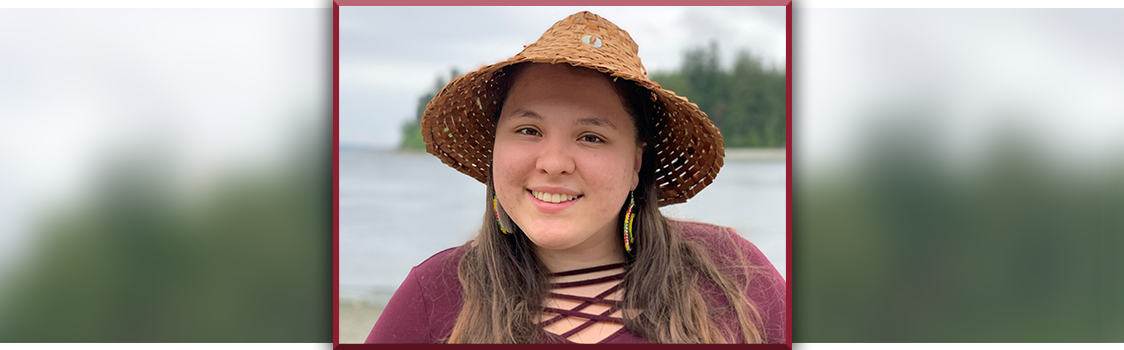 Hailey Crow wearing a woven hat and a burgundy top, standing near a lakeshore with trees along the distant shoreline.