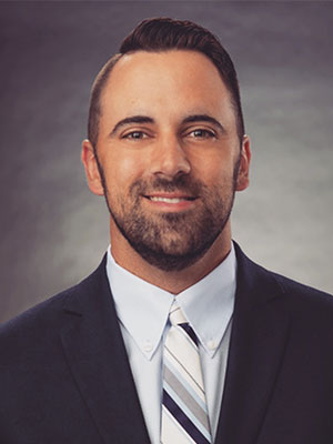 Jay Harris wearing a dark suit jacket, white shirt, and striped tie, posed against a softly lit gray background.