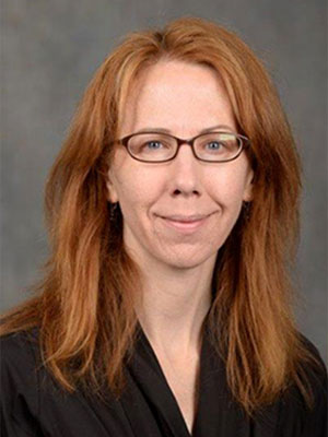 Kristen Kuhn wearing a black top, with shoulder-length red hair, posed against a neutral gray background.