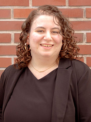 Lydia Guenther wearing a black top and blazer with curly hair, standing in front of a red brick wall.