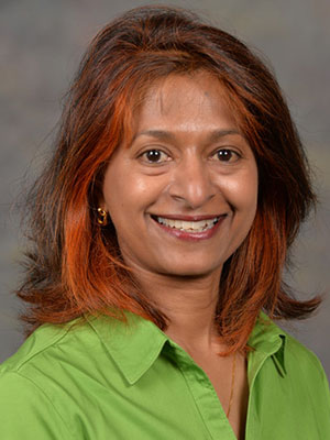 Minie Yadama wearing a bright green collared shirt, with shoulder-length brown hair highlighted with copper tones, posed against a neutral gray backdrop.