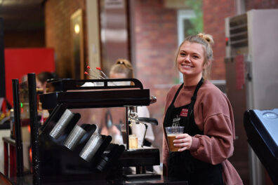 A barista in a café holds an iced drink while standing beside an espresso machine and cup dispensers.