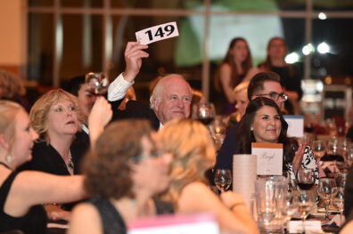 Guests seated at a formal event table as one attendee raises a bidding paddle labeled “149” during an auction.