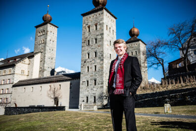 A person wearing a dark suit and a WSU scarf stands outdoors in front of historic stone towers with onion-shaped domes against a clear blue sky.