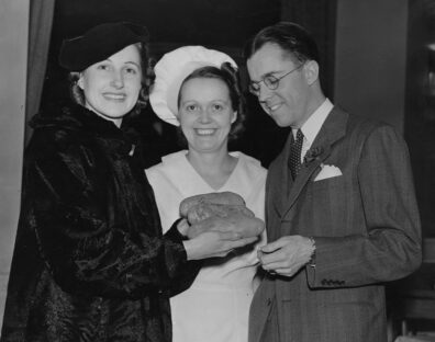 Three people stand together in a vintage scene as one of them presents a loaf of bread to another, with a chef’s hat visible on the person in the middle.