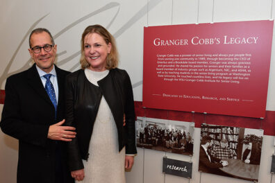 A man and woman stand together in front of a display titled ‘Granger Cobb’s Legacy,’ featuring text panels and historic photos related to the senior living pioneer.