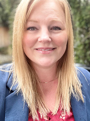 Rachel Boyer wearing a blue blazer over a red top, standing outdoors with greenery softly blurred in the background.