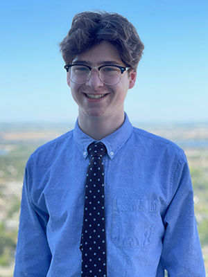 Ryan McKinley standing outdoors in a blue button‑down shirt and polka‑dot tie with a clear sky and landscape in the background.