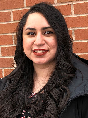 Tabetha Villanueva wearing a black jacket over a floral top, standing in front of a red brick wall.