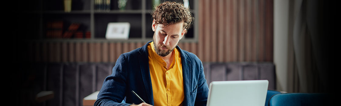 A person working at a laptop in a modern office space, holding a pen and wearing a yellow shirt with a dark jacket.