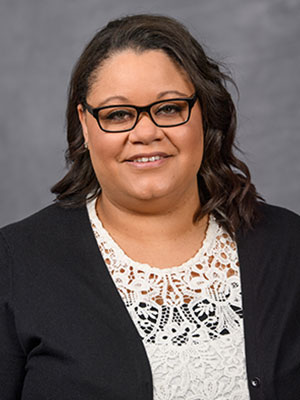 Whitney Thurgood wearing a black cardigan over a white lace top, posed against a gray studio background.