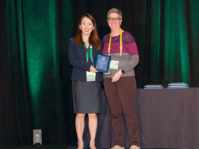 Yafang Li and Associate Professor Michelle Carter stand onstage holding the 2021 Doctoral Student Service Award plaque against a dark green backdrop.