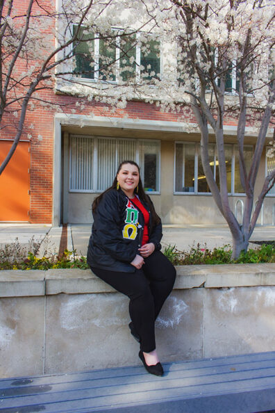 Hailey Crow of Alpha Pi Omega Sorority Inc. sitting on a stone ledge beneath blooming trees outside a brick campus building.