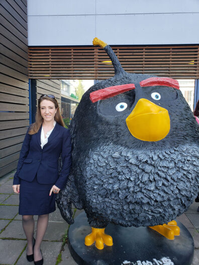 Melanie Wood in business attire stands beside a large Angry Birds character statue outside Rovio Entertainment’s building