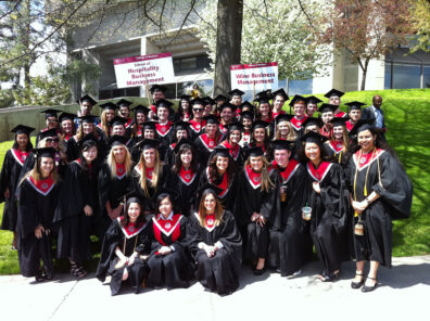A large group of graduates in black caps and gowns pose outdoors on a sunny day, standing beneath signs for Hospitality Business Management and Wine Business Management.