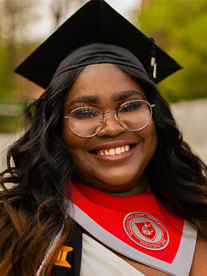 Perebotie Amughan wearing a graduation cap and a red WSU stole, standing outdoors with greenery in the background.