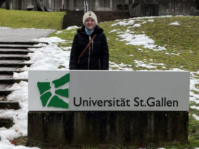 Nicole Dalton stands outdoors in winter clothing behind a Universität St. Gallen sign, with snow patches and steps in the background.