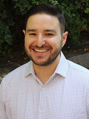 Alex Kier standing outdoors in a light checkered shirt with greenery in the background.