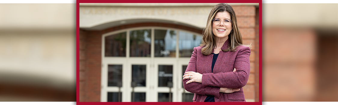 Cheryl Oliver standing with arms crossed in front of a building entrance, wearing a burgundy blazer and dark top.