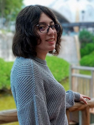 Claudia Jacobo standing outdoors by a wooden railing, wearing a gray knit sweater with greenery in the background.