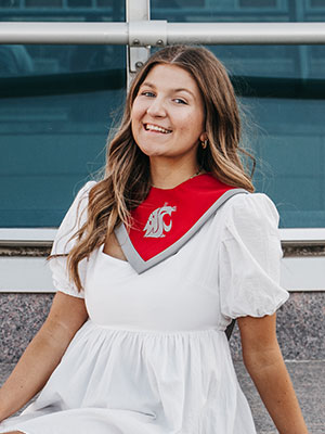 Katie Vail wearing a white dress and a crimson and gray WSU stole while sitting outdoors in front of a modern glass building.
