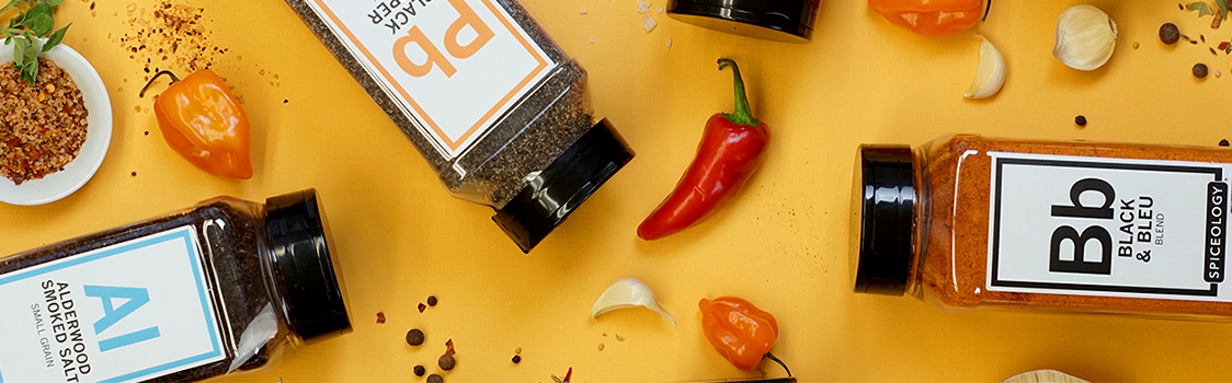 Spiceology seasoning bottles arranged on a yellow surface among peppers, garlic cloves, peppercorns, and spices.
