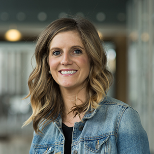 Keri Rhodes wearing a denim jacket over a black top, standing indoors with soft lights and blurred hallway details in the background.
