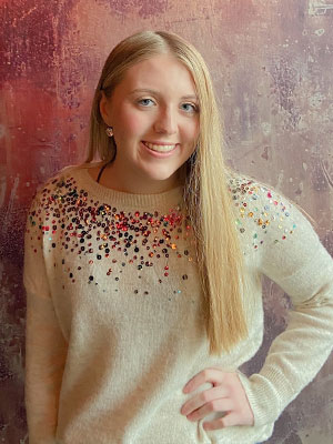 Molly Williams standing against a textured reddish‑purple backdrop, wearing a cream sweater decorated with colorful sequins across the neckline.