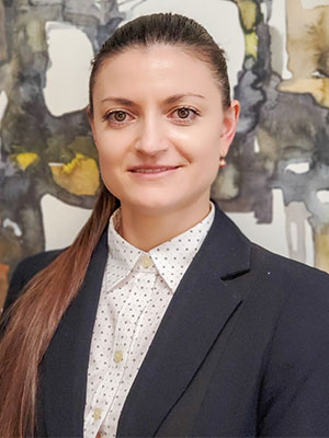 Olga Gira wearing a dark blazer and patterned button‑up shirt, standing in front of an abstract painted background.