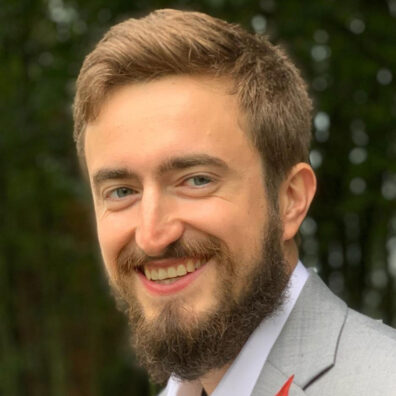 Sean Russell wearing a light gray suit jacket and white shirt, pictured outdoors with green foliage in the background.