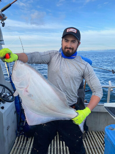 Sean Russell standing on a boat and holding a large fish with both hands against an ocean backdrop.