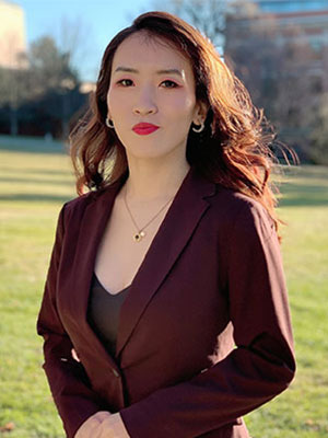 Yafang Li standing outdoors in a burgundy blazer on a sunny lawn with trees and a building in the background.