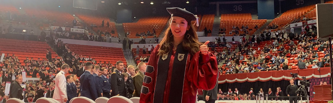Yafang Li standing in graduation regalia at a large indoor ceremony, giving a thumbs‑up amid rows of seats and a crowd in the arena.