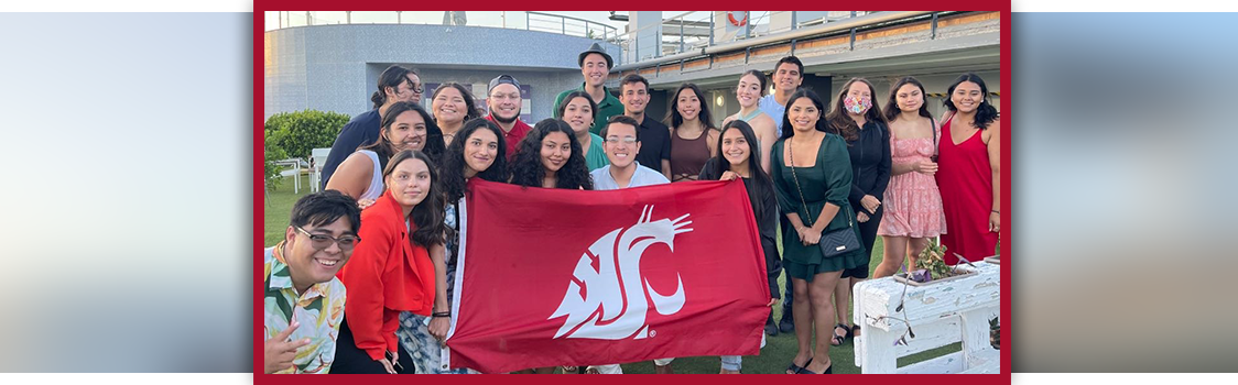Group of students gathered outdoors holding a large crimson WSU flag, posing together in front of a campus-style building.