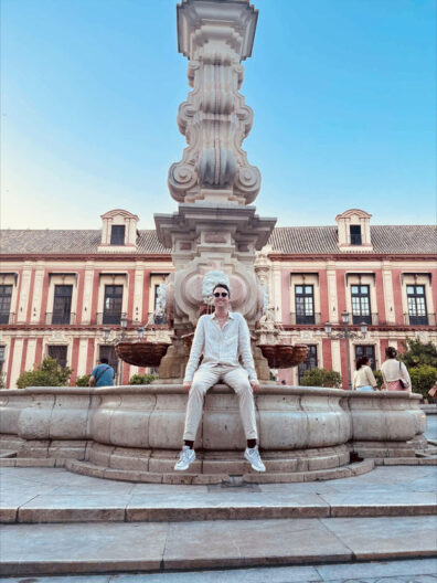 Kole Lappe sits on the edge of an ornate stone fountain in a historic plaza, with a tall sculpted centerpiece and colorful building façades behind him.
