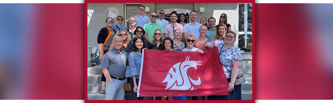 OMBA students and faculty pose together on outdoor steps in Prague, holding a large crimson WSU flag in front of a building.