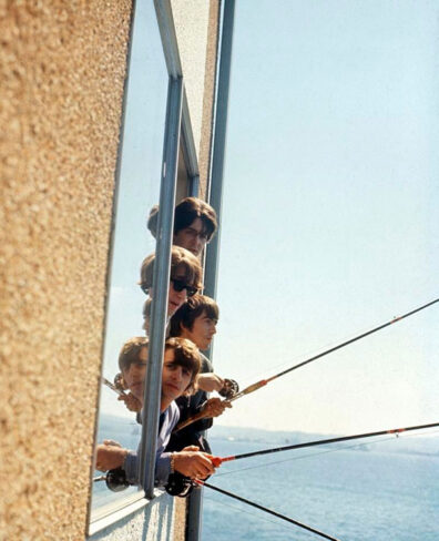 Members of The Beatles lean out of a hotel window at the Edgewater in 1964, holding fishing rods out over the water on a bright, clear day.