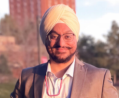 Gurdeep Singh Raina standing outdoors in warm sunlight, wearing a light-colored turban, suit jacket, and collared shirt with a red beaded accessory around the neck.