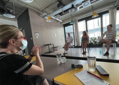 Students listen to a panel of entrepreneurs at HubHub Prague, with three speakers seated on a small stage and notebooks, drinks, and tables visible in the foreground.