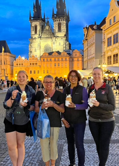 MBA students enjoying chimney bread and ice cream in Prague Square at dusk, standing before the illuminated Church of Our Lady before Týn and surrounding historic buildings.